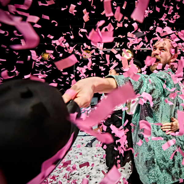 a dancer in a sequin jacket holds out a bowler hat towards the camera and grins widely as confetti rains down