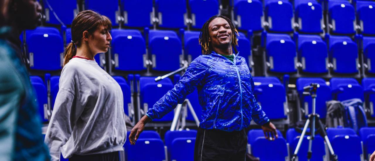 Freelance dancer Anthony Matsena stretches in a dance studio in front of blue seating with female dancer Nikita 