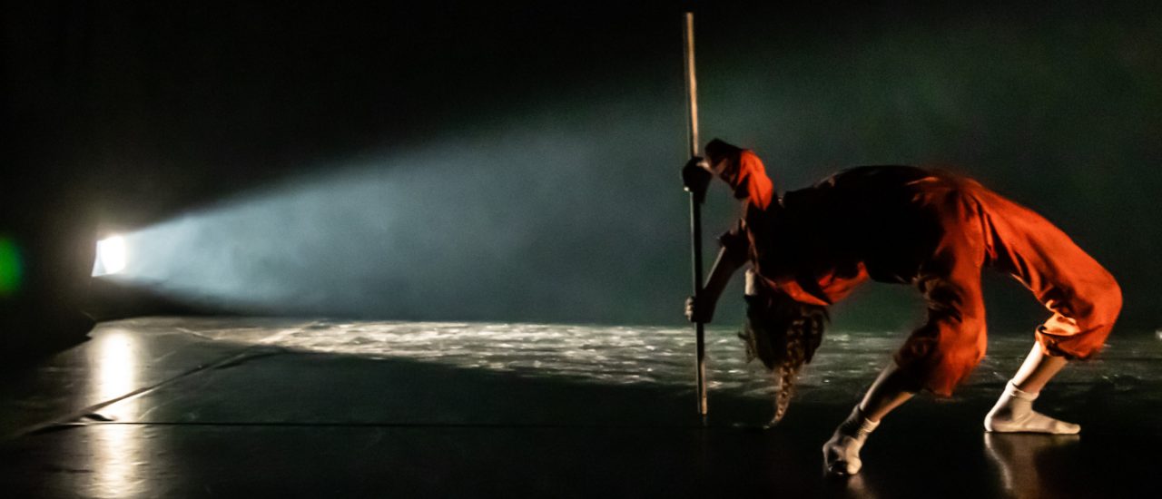 A dancer in an orange jumpsuit leans backwards in an arch