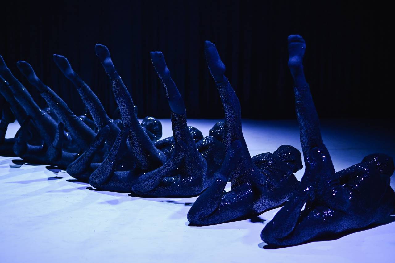 a line of dancers in black glitter costumes linked together by their legs