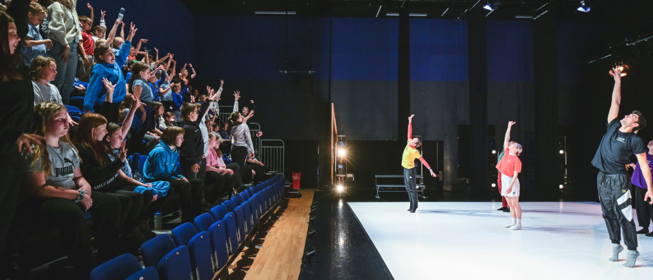 dancers on stage leading a school in a warm up, each child is smiling and raising their arms. 