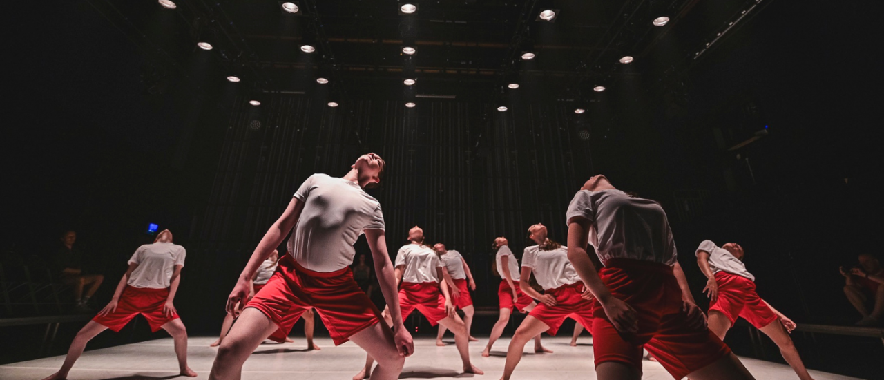 a number of young dancers in white t-shirts and red sports shirts leaning back under rows of spotlights