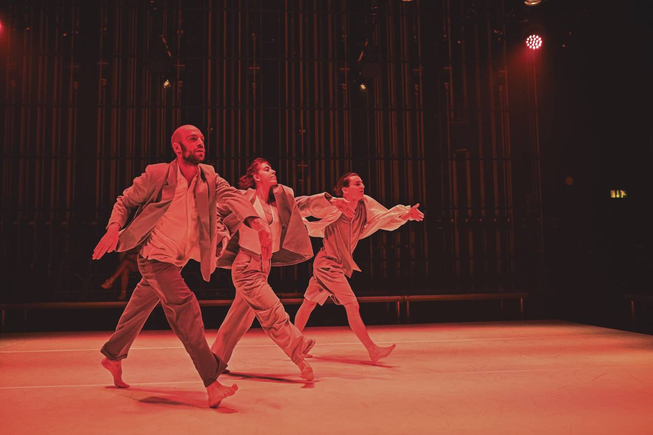 three dancers stride across the stage in time, arms out swinging