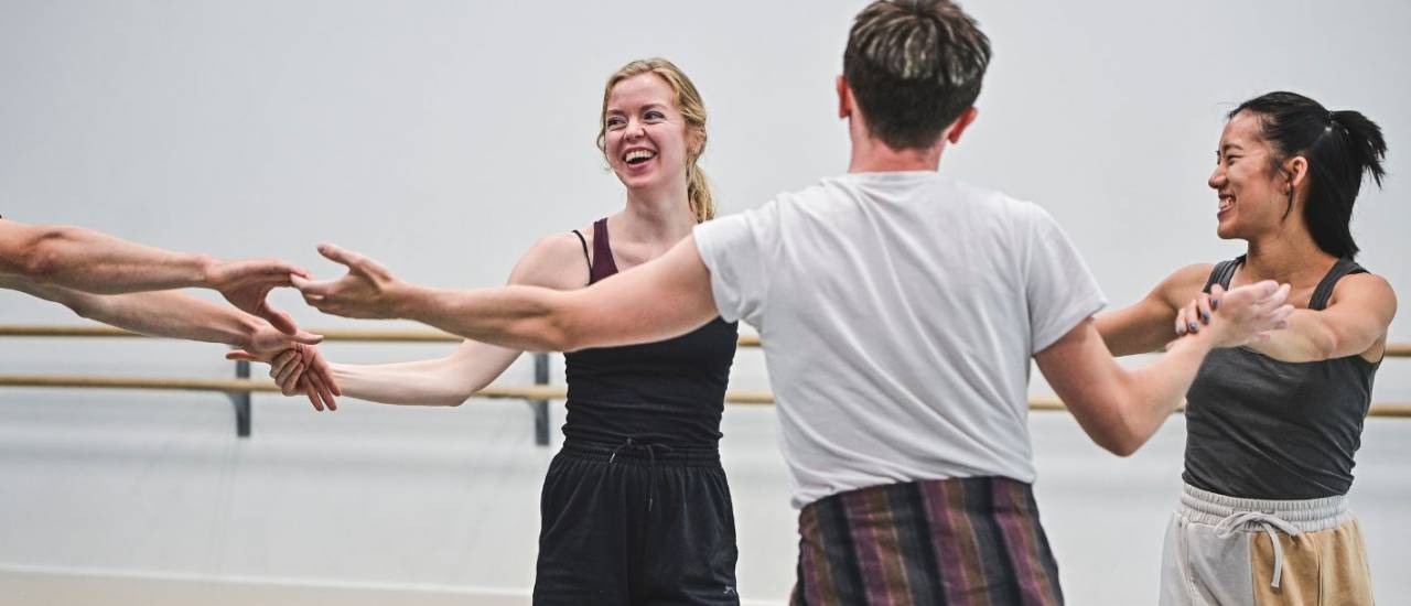 three dancers in casual clothes and one out of frame hold hands in a circle and are grinning at one another