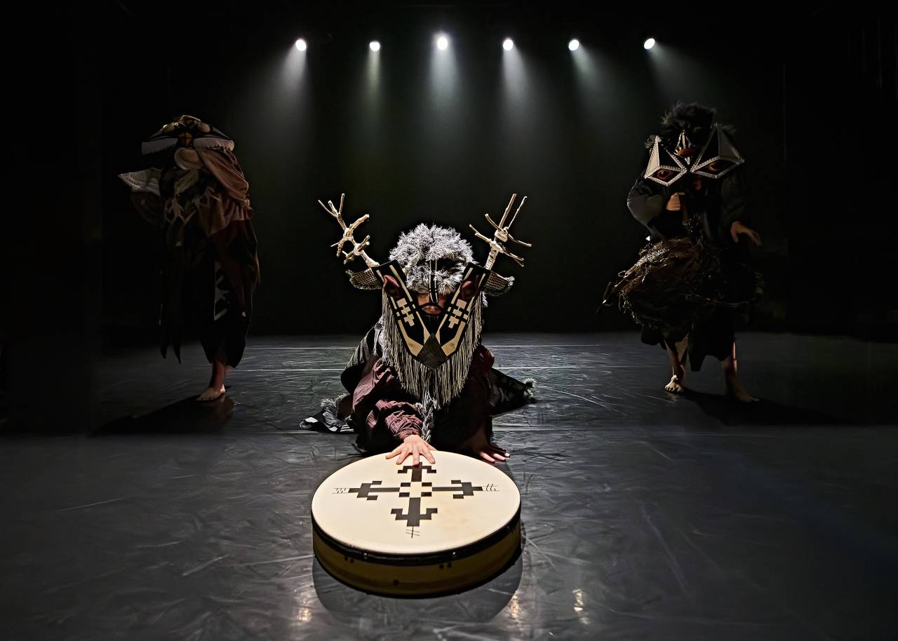 a dramatic scene, two dancers blend into the background whilst one in a stag mask with horns made of sticks looks directly at the camera, sliding a drum across the floor  