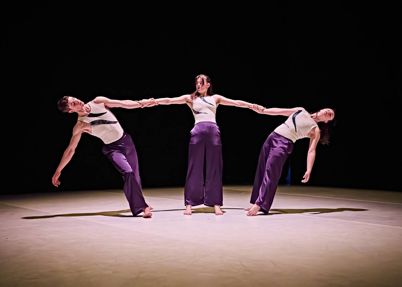 riz stands at the centre of a spot light, arms out counter balancing two other dancers