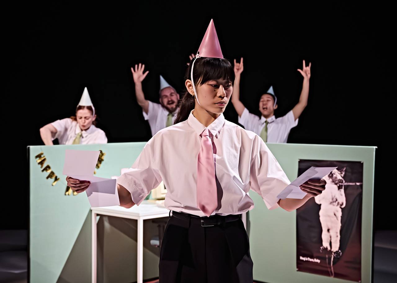 Jill holds two pices of paper and wears a shirt, tie and party hat - she looks like she's making a decision whilst behind her other dancers have a new years eve party