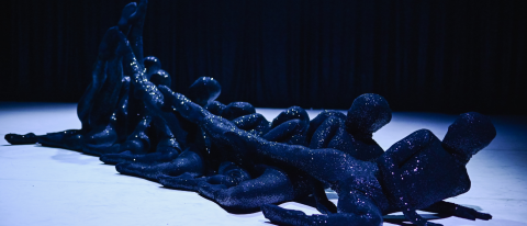 dancers in black sequin jumpsuits and masks in a line with parallel limbs