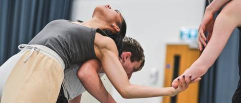 one dancer leans over whilst another backbends over them, weight supported on their backm and holding the hand of another