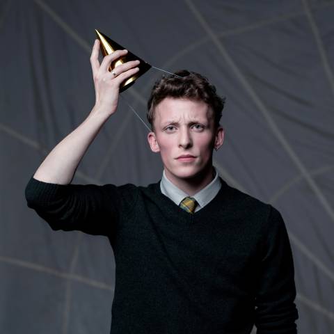 John-William wears a black jumper, white shirt and gold tie, and a confused expression as they pull a party hat strapped under their chin with elastic away from their head, they have short, softly curled hair and rosy cheeks