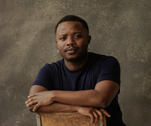 Bakani Pick-Up has short cropped hair and sits backwards on a wooden chair with a serious expression