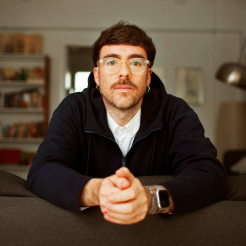Marcos Morau has short brown hair and a neat moustache and stubble, and trendy see-through framed glasses, he leans forward towards the camera slightly, hands clasped infront of him on a table